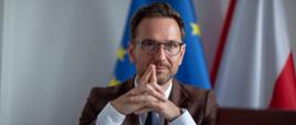 Minister Waldemar Buda siedzi przy stole konferencyjnym, za nim flagi Polski i UE
