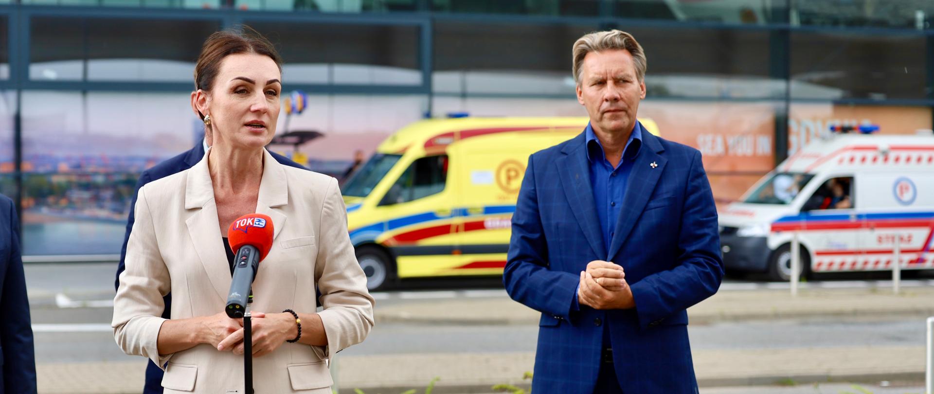 kobieta przemawia przed terminalem na lotnisku, w tle widać karetki pogotowia