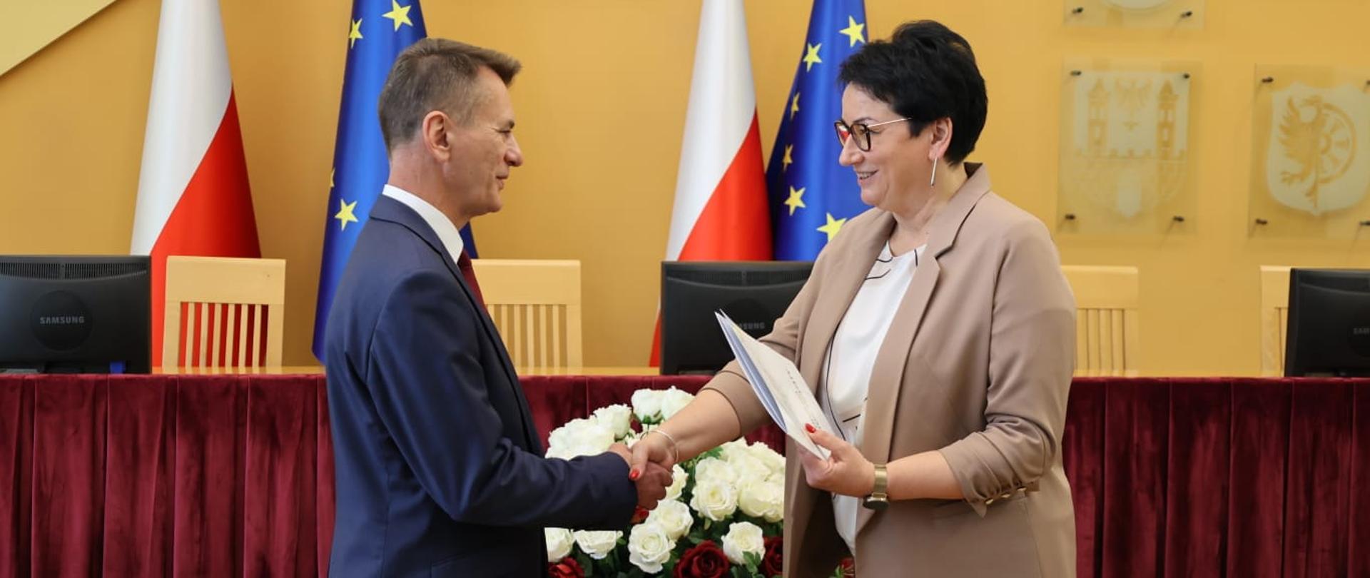 Wojewoda Opolski Monika Jurek wręcza powołanie nowemu wicekuratorowi oświaty, Markowi Leśniakowi. Na zdjęciu widać ich uścisk dłoni. Oboje się uśmiechają. W tle widać flagi Polski i Unii Europejskiej. 