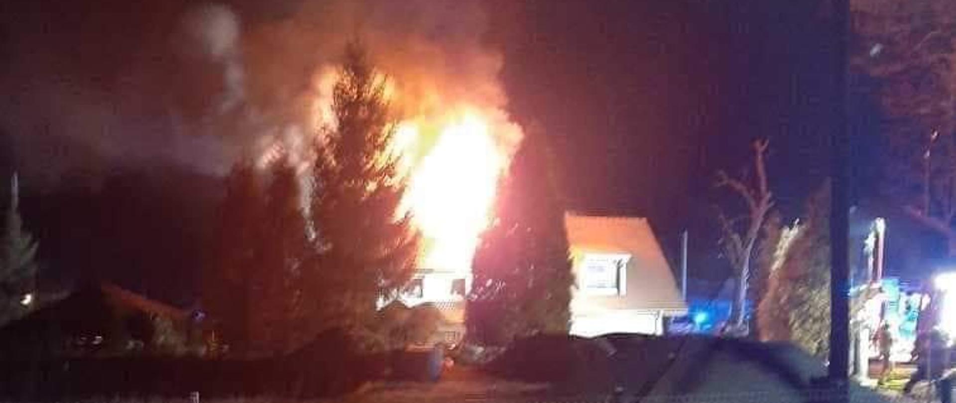 Obraz przedstawia płonący budynek mieszkalny. Płomienie wychodzą ponad dach budynku. Pora nocna.