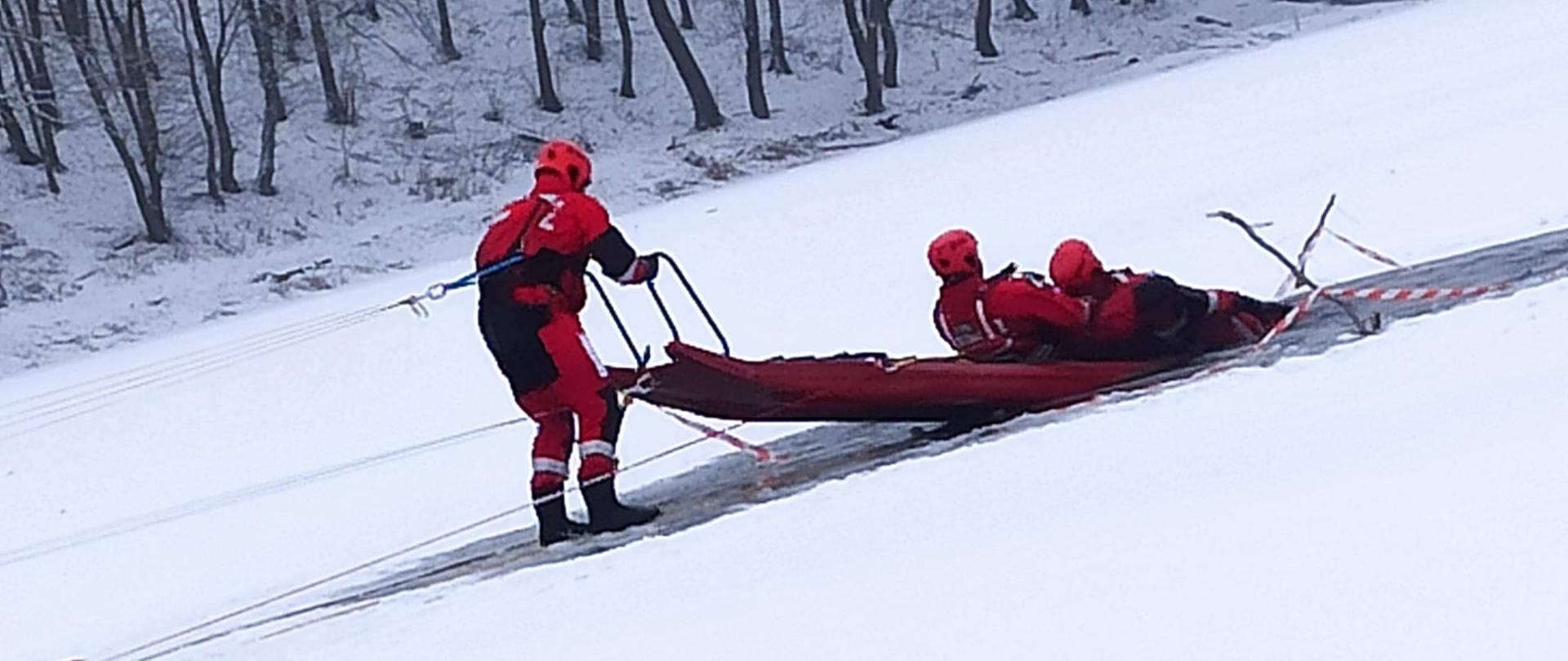 Trzech ratowników ubranych w czerwone kombinezony i kaski, prawdopodobnie strażaków, wykonuje ćwiczenia ratownicze na stoku pokrytym śniegiem. Jeden z ratowników stoi, ciągnąc za sobą czerwne nosze, na których leżą dwaj inni ratownicy. Cała scena odbywa się na pochyłym, zaśnieżonym terenie. W tle widoczny jest las z nagimi drzewami, a horyzont jest zasłonięty przez śnieżycę lub zamglenie.