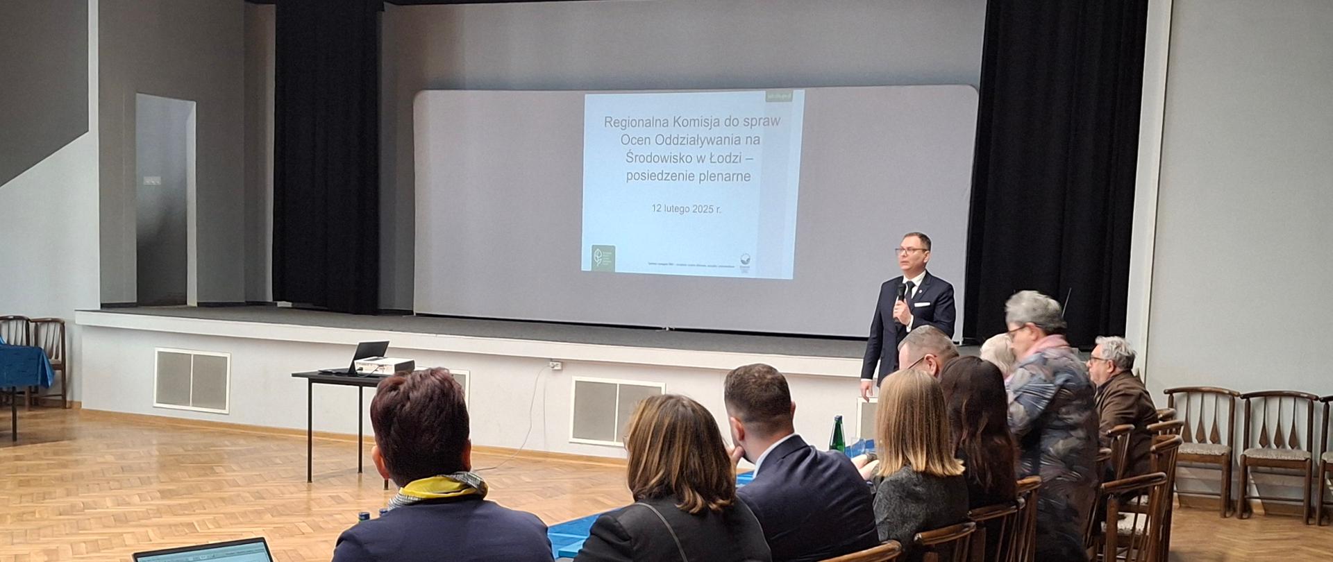 Sala, na ścianie wyświetlony slajd z napisem Komisja do spraw Ocen Oddziaływania na Środowisko - posiedzenie plenarne 12 lutego 2025 r. Mężczyzna w garniturze z mikrofonem stroi zwrócony przodem do ludzi siedzących przy stole.