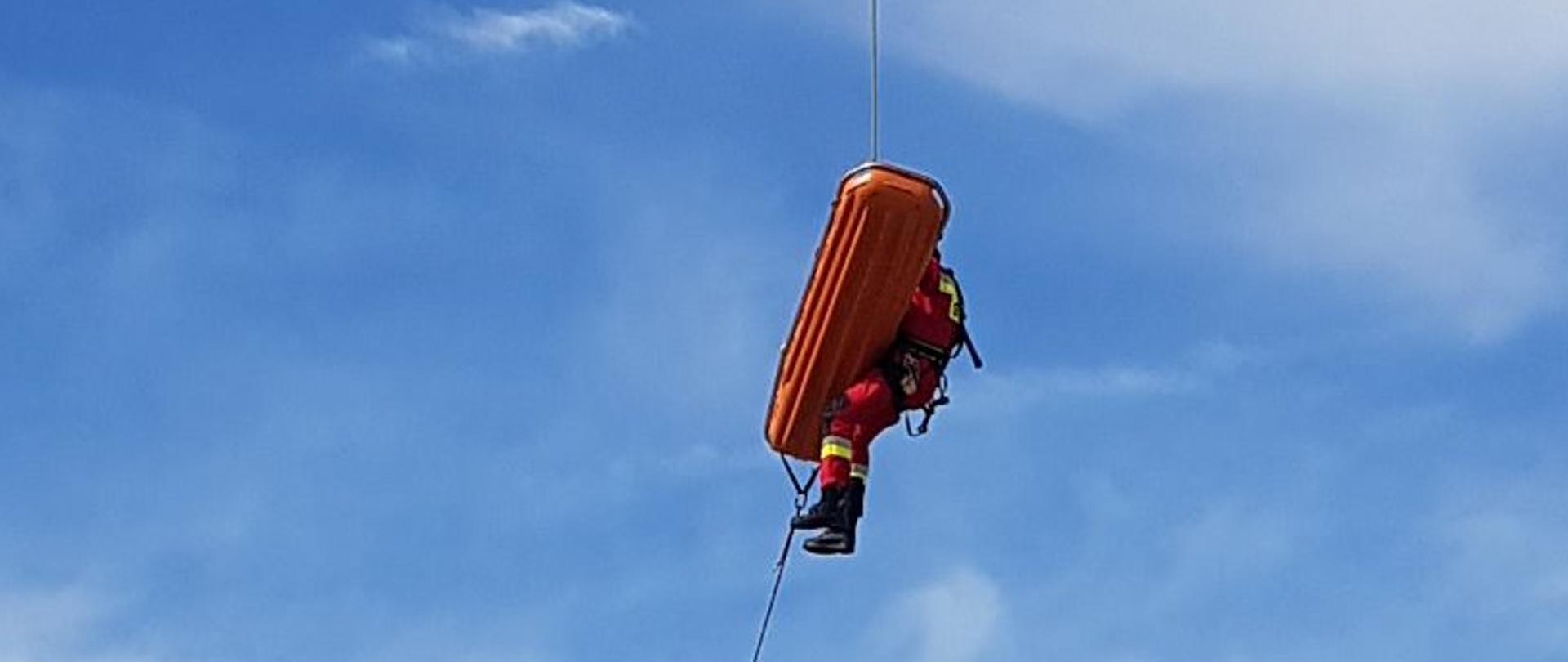 Grupa strażaków stoi na ziemi i asekuruje strażaka zjeżdżającego na linie z doczepionymi noszami.