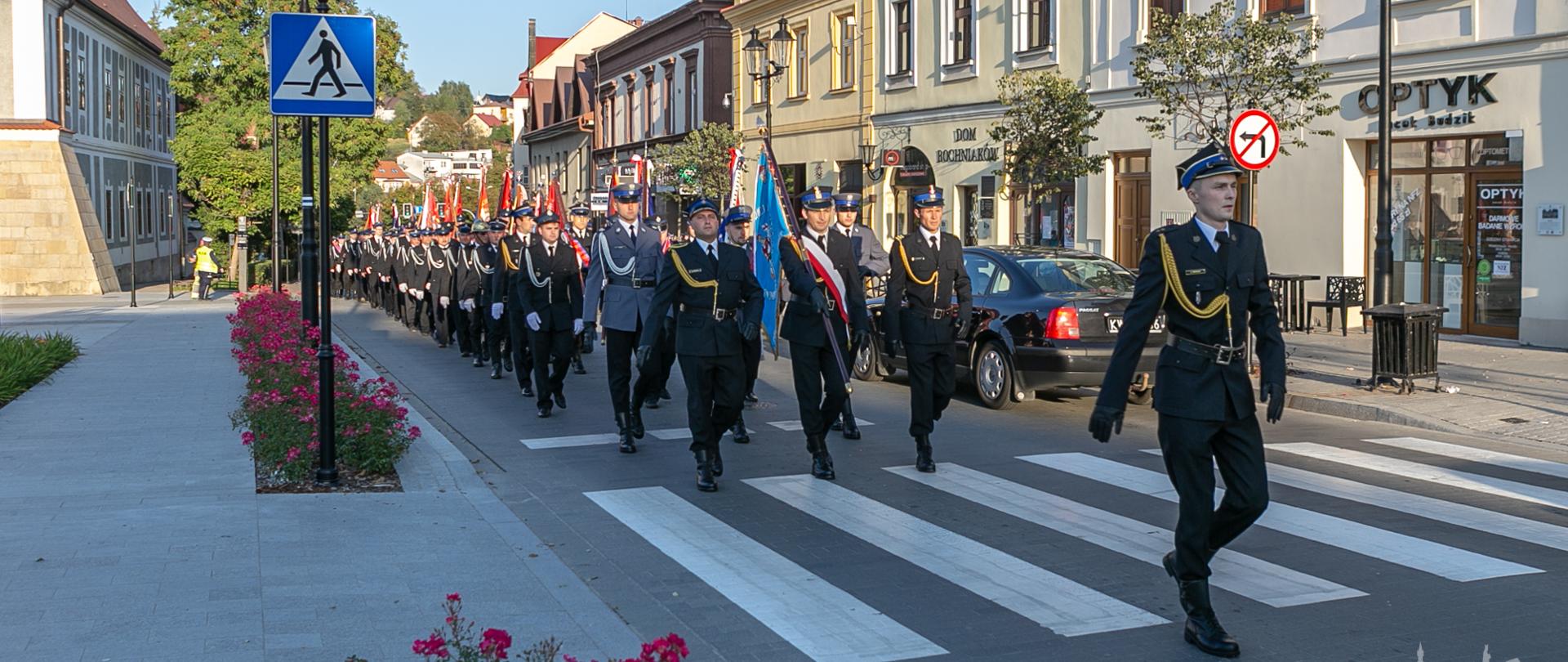 Na pierwszym planie strażak w mundurze wyjściowym, który jako Dowódca prowadzi oddział strażaków Państwowej i Ochotniczej Straży Pożarnej ze sztandarami. W tle ulica i zabudowa centrum miasta Bochni z charakterystycznymi kamienicami. 