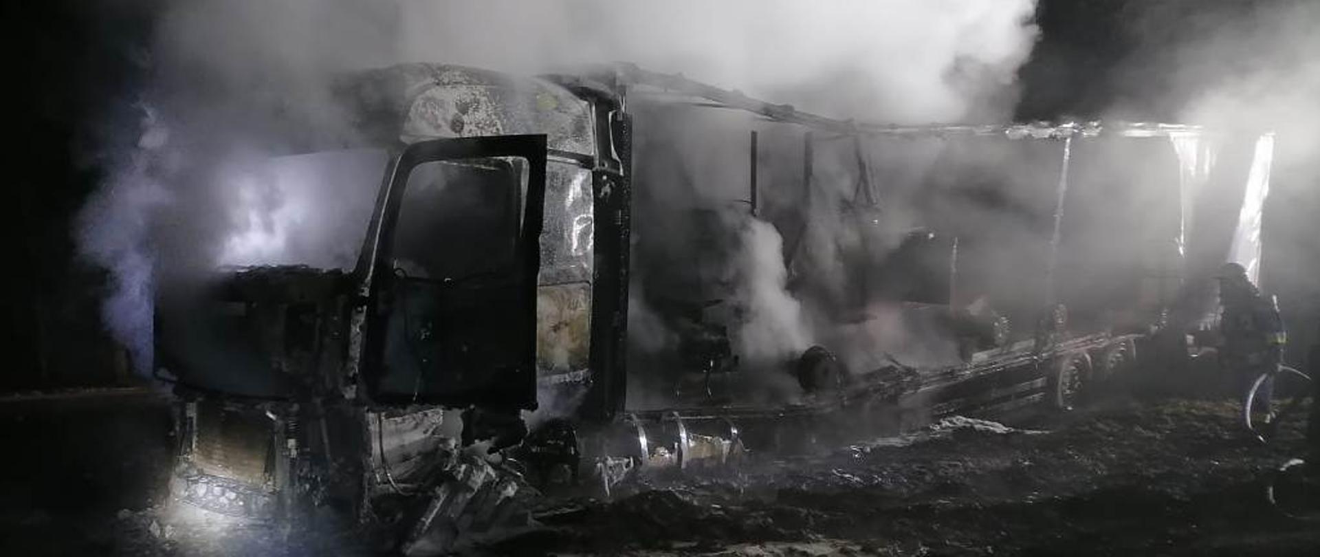 Ciągnik siodłowy z naczepą, który uległ praktycznie całkowitemu spaleniu. Zarówno z kabiny jak i z naczepy unoszą się kłęby pary po podjętych przez strażaków działaniach gaśniczych.