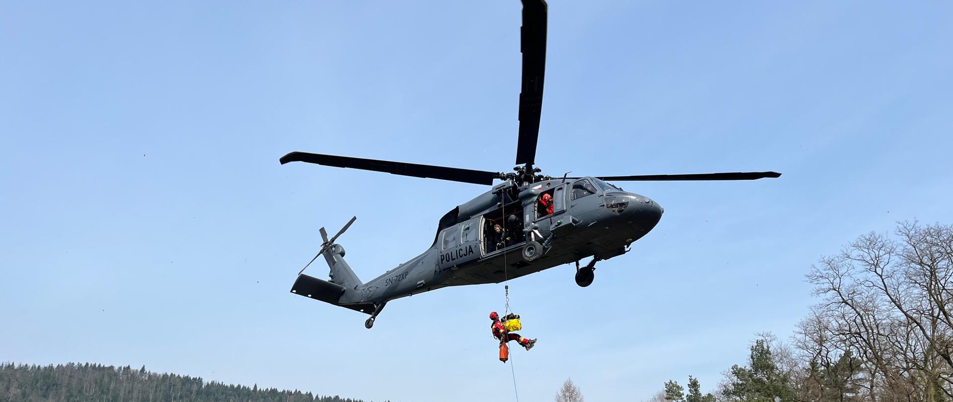 Zdjęcie przedstawia strażaka z noszami opuszczonego na linie z lecącego śmigłowca. W tle widoczny las oraz stacja energetyczna.