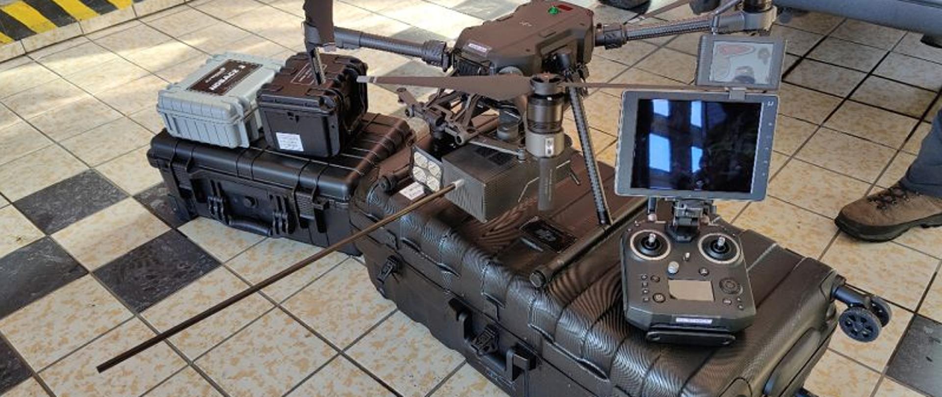 Zdjęcie przedstawia statek bezzałogowy – dron Matrice 210. Urządzenie stoi na walizkach w pomieszczeniu garażowym. Podłoga pokryta szarymi płytkami gresowymi. Z prawej strony konsola sterująca, z lewej czterowirnikowy dron.