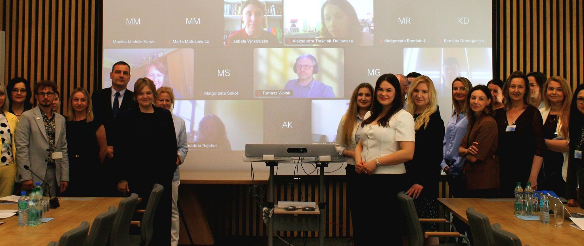 Przed wbudowanym w ścianę z cienkich pionowych listewek ekranem na którym widać kafelki z twarzami osób łączących się zdalnie stoją po obu stronach ekranu dwie kilkunastoosobowe grupy ludzi, z lewej wiceminister Zioło-Pużuk.