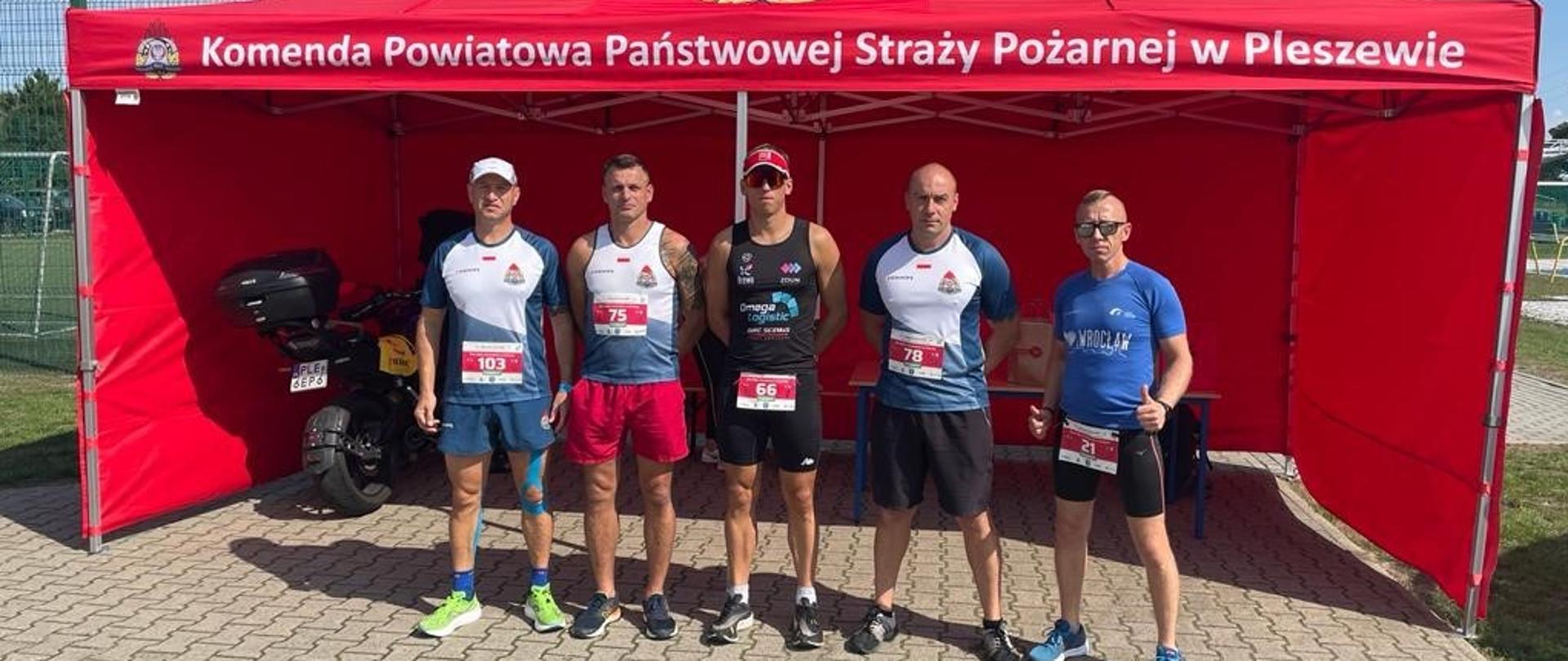Czterech ubranych na sportowo mężczyzn stoi obok siebie na drodze asfaltowej, w tle widoczny czerwony namiot z napisem Komenda Powiatowa Państwowej Straży Pożarnej w Pleszewie