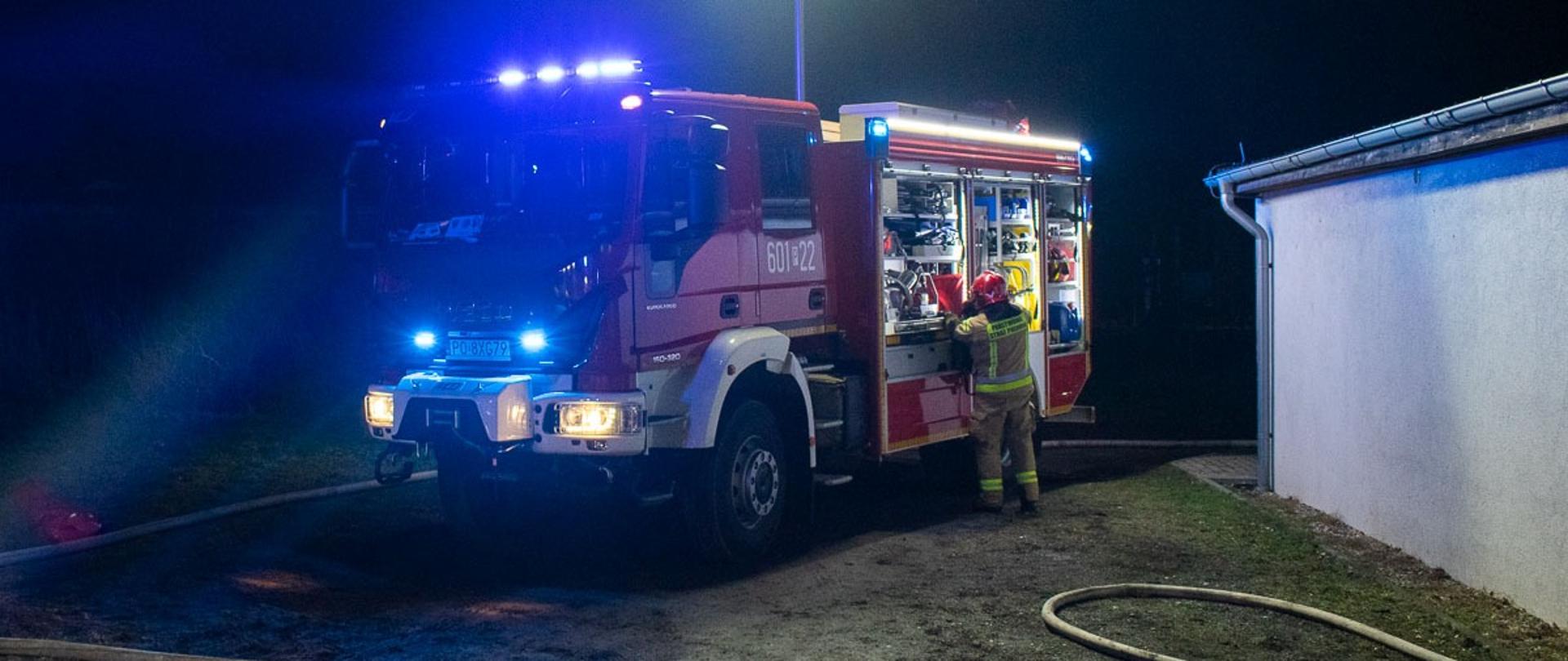 Na zdjęciu czerwony samochód strażacki z włączonymi sygnałami świetlnymi koloru niebieskimi, obok samochodu strażak w ubraniu koloru żółtego i czerwonym hełmie.