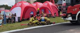 Strażacy podczas ćwiczenia ratowniczego. W tle namioty wystawowe z napisami STRAŻ, samochód pożarniczy oraz dźwig. 