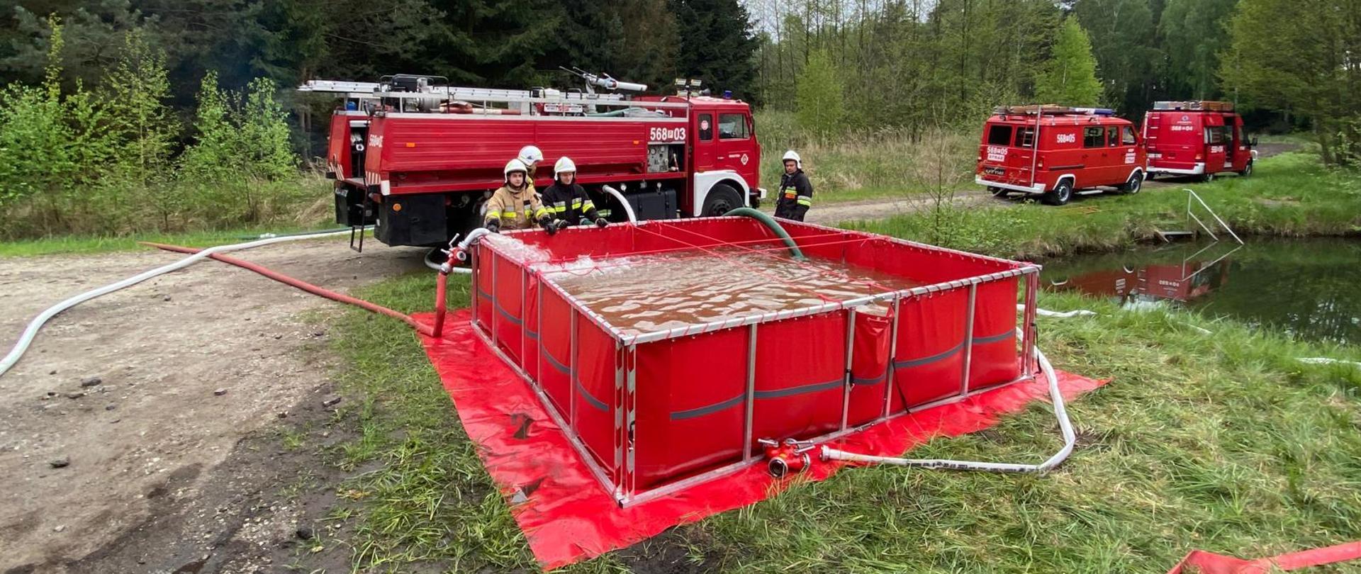 na leśnej drodze stoją trzy samochody pożarnicze, przed jednym duży czerwony zbiornik z wodą i wężami , przy zbiorniku stoją strażacy