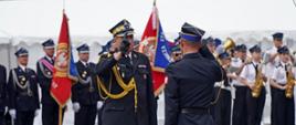 Na zdjęciu widoczni strażacy Państwowej Straży Pożarnej przyjmujący meldunek Małopolski Komendant Wojewódzki i składający meldunek oficer w tle widoczne poczty sztandarowe z jednostek Ochotniczych Straży Pożarnych i orkiestra dęta.