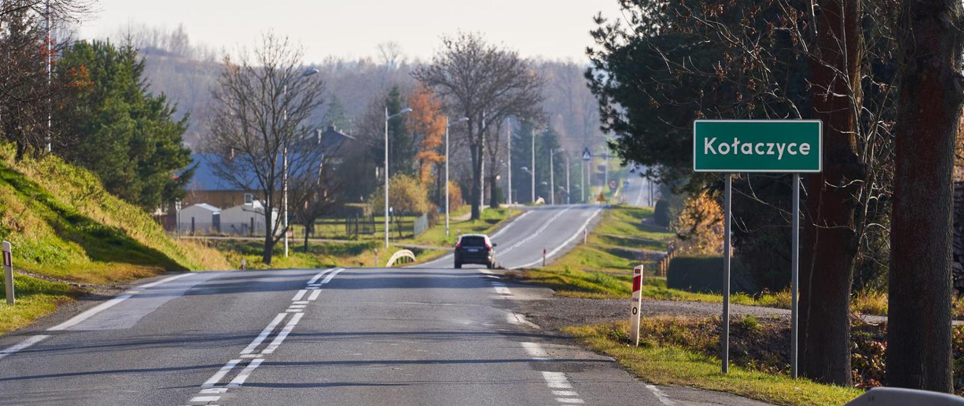 Znak z nazwą miejscowości „Kołaczyce” ustawiony przy drodze przebiegającej przez teren zabudowany. Na jezdni widoczny jest samochód jadący w kierunku centrum miejscowości. Po obu stronach drogi znajdują się drzewa, pojedyncze budynki oraz latarnie uliczne. Zdjęcie wykonano w pogodny dzień.