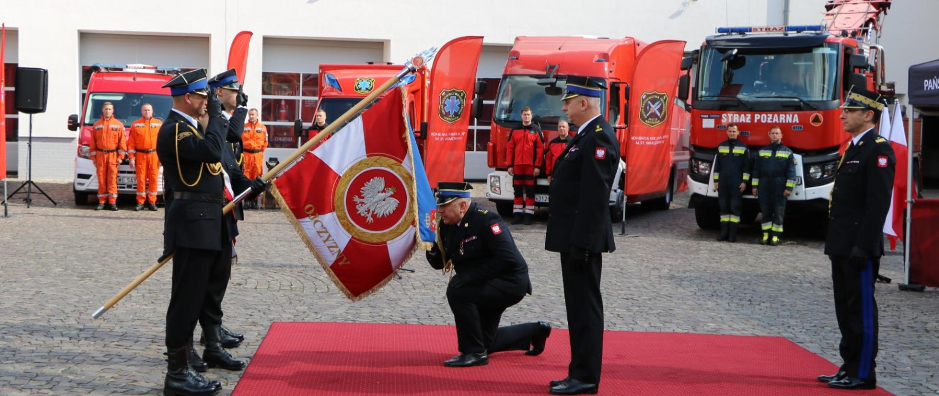 Komendant zdający obowiązki klęczy na czerwonym dywanie przed sztandarem z orłem i dwiema innymi osobami ubranymi w galowe mundury strażackie. W tle widać strażaków w pomarańczowych i granatowych mundurach oraz kilka czerwonych wozów strażackich. Na flagach za pojazdami widnieje godło Państwowej Straży Pożarnej. Ujęcie zostało zrobione w ciągu dnia na brukowanym dziedzińcu. 