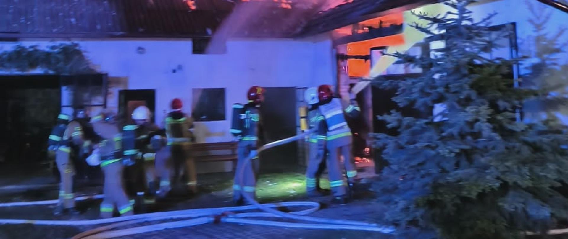 Zdjęcie z miejsca pożaru. Pora nocna. Widać budynek gospodarczy objęty pożarem. W części poddasza i dachu widoczne płomienie oraz dym. Strażacy stoją przed budynkiem i podają prądy gaśnicze na obiekt objęty pożarem. Strażacy ubrani są piaskowe ubrania oraz hełmy. Kilu strażaków ma na sobie aparaty powietrzne służące ochronie dróg oddechowych przed oddziaływaniem dymu i innych czynników szkodliwych. Jeden ze strażaków posiada dodatkowo biało-niebieską kamizelkę oznaczającego kierującego działaniem ratowniczym. W utwardzonym podłożu widać rozwinięte węże strażackie. 