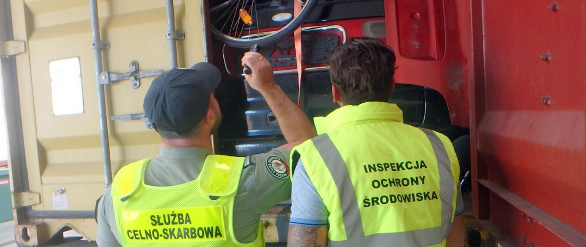 W pozycji stojącej dwóch mężczyzn obróconych tyłem ubranych w kamizelki (jeden z nich ma podniesioną prawą rękę trzymając latarkę) kierują swój wzrok na ciężarówkę na której są otwarte tylne drzwi, a w niej różne przedmioty.