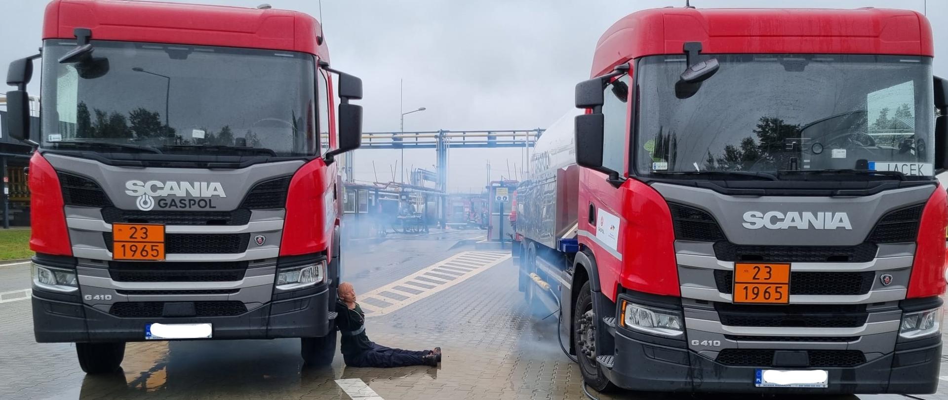 Zdjęcie przedstawia dwie autocysterny znajdujące się na terenie zakładu Gaspol. Koło jednej z nich ułożony jest manekin. Spod drugiej wydobywa się zadymienie.