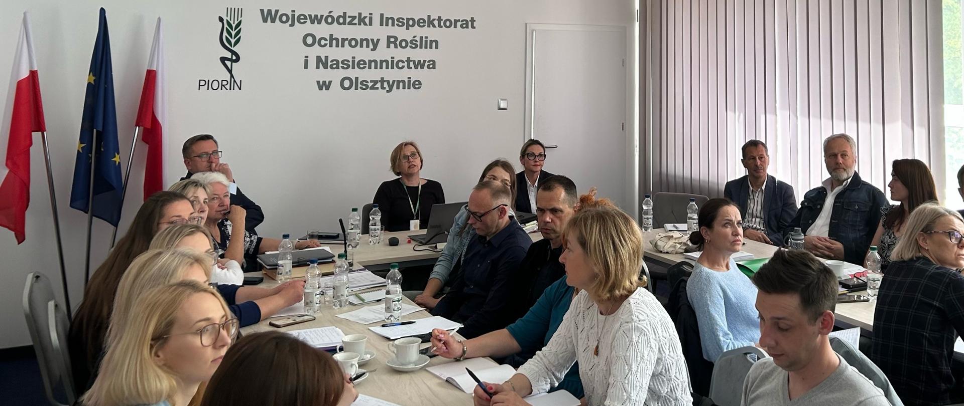 : : Przy stołach ustawionych w literę C siedzą osoby, siedząca na środku kobieta ma przed sobą
laptopa, uczestnicy zwróceni są twarzą do przeciwległej ściany