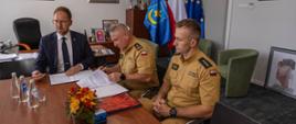W pomieszczeniu z szarymi ścianami przy stole siedzą: prezydent miasta ubrany w granatowy garnitur, komendant w piaskowym mundurze, oraz drugi strażak również w piaskowym mundurze. Na stole leżą dokumenty. W tle stojak z flagami.