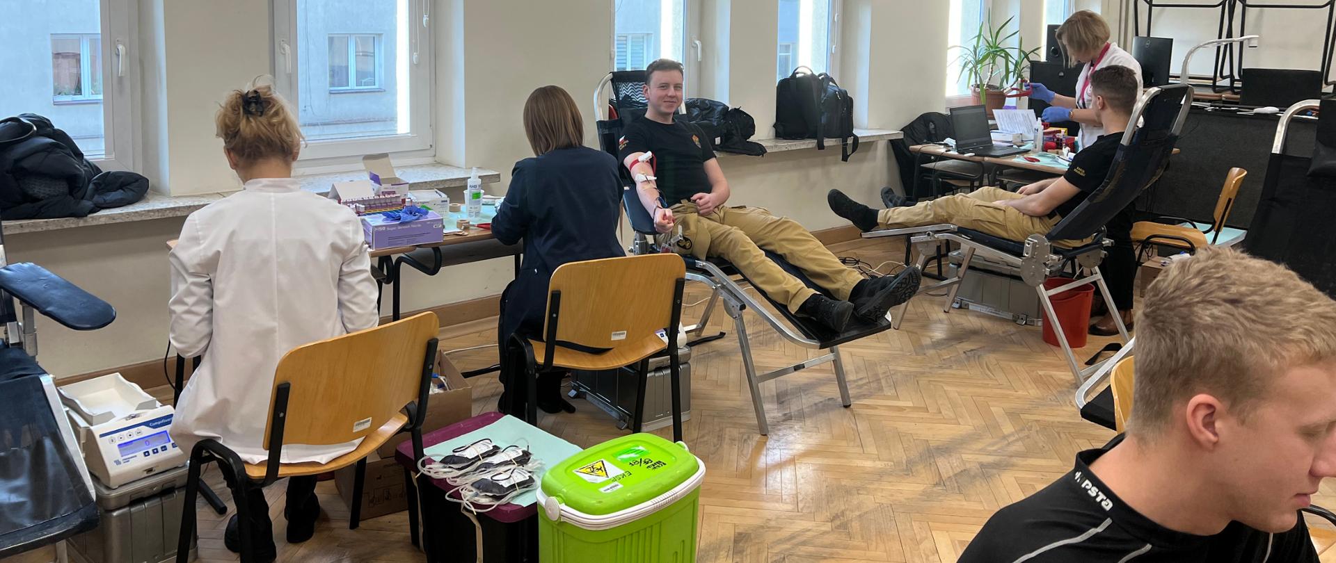 Wnętrze sali lekcyjnej. Na środku ustawione stoliki i przyrządy do pobierania krwi, w tym dwa fotele na których siedzi dwóch mężczyzn. W sali obecne są także inne osoby uczestniczące w donacji.