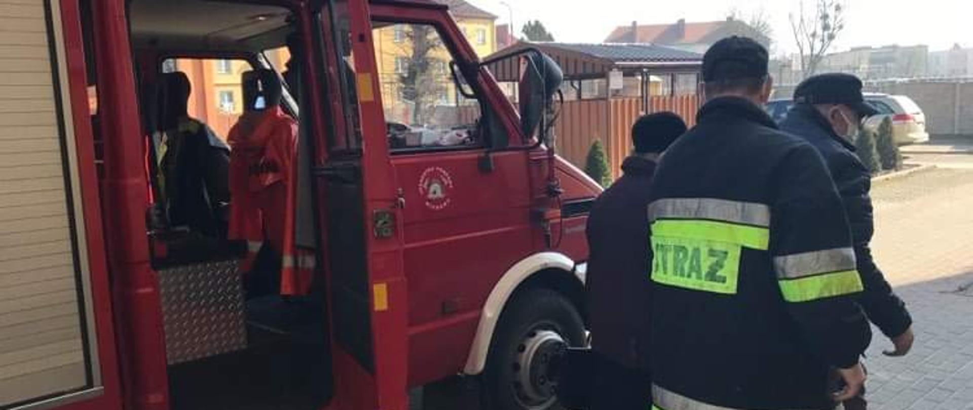 Pochmurnie. Strażak w ciemnym ubraniu pomaga wysiąść z czerwonego samochodu pożarniczego dwóm starszym osobom.