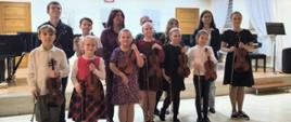 Dzieci stoją ze skrzypcami pod sceną wraz z nauczycielką skrzypiec i akompaniatorką pozując do wspólnego zdjęcia po zakończonej audycji. 