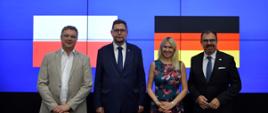 Przed wbudowanym w ścianę z cienkich pionowych listewek wielkim ekranem na którym widać flagi Polski i Niemiec na niebieskim tle stoi mężczyzna w jasnoszarym garniturze, wiceminister Szeptycki, kobieta w kolorowej sukience i mężczyzna w czarnym garniturze.
