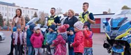 grupka dzieci wraz z opiekunkami i policjantami pozują do pamiątkowego zdjęcia z motorami za nimi znajdują się garaże na samochody strażackie