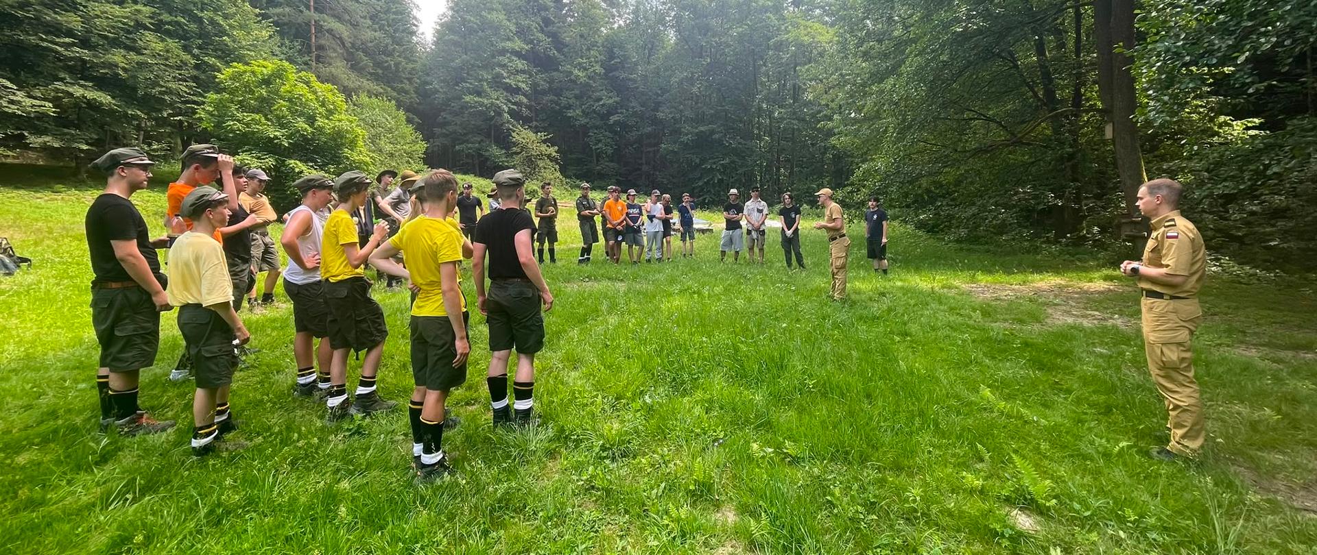 Na zielonej łące stoi strażak w piaskowym mundurze, naprzeciw niego grupa harcerzy w krótkich spodenkach i podkoszulkach na głowach mają rogatywki. Dookoła drzewa.