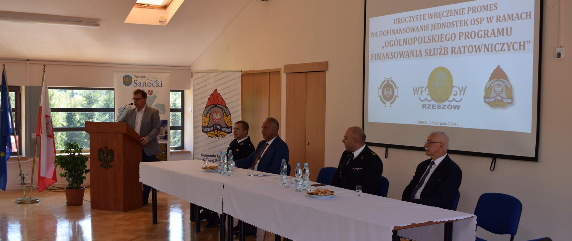 Zdjęcie zrobione w pomieszczeniu świetlicy KP PSP Sanok. Na zdjęciu znajdują się 4 osoby w pozycji siedzącej przy stole: komendant powiatowy PSP w Sanoku bryg. Grzegorz Oleniacz, prezes WFOŚiGW w Rzeszowie Adam Skiba, wicestarosta sanocki Janusz Cecuła i prezes Zarządu Oddziału Powiatowego Związku Ochotniczych Straży Grzegorz Klecha. Po prawej stronie siedzących osób znajduje sie mównica przy której stoi i przemawia poseł na sejm RM Piotr Babinetz.