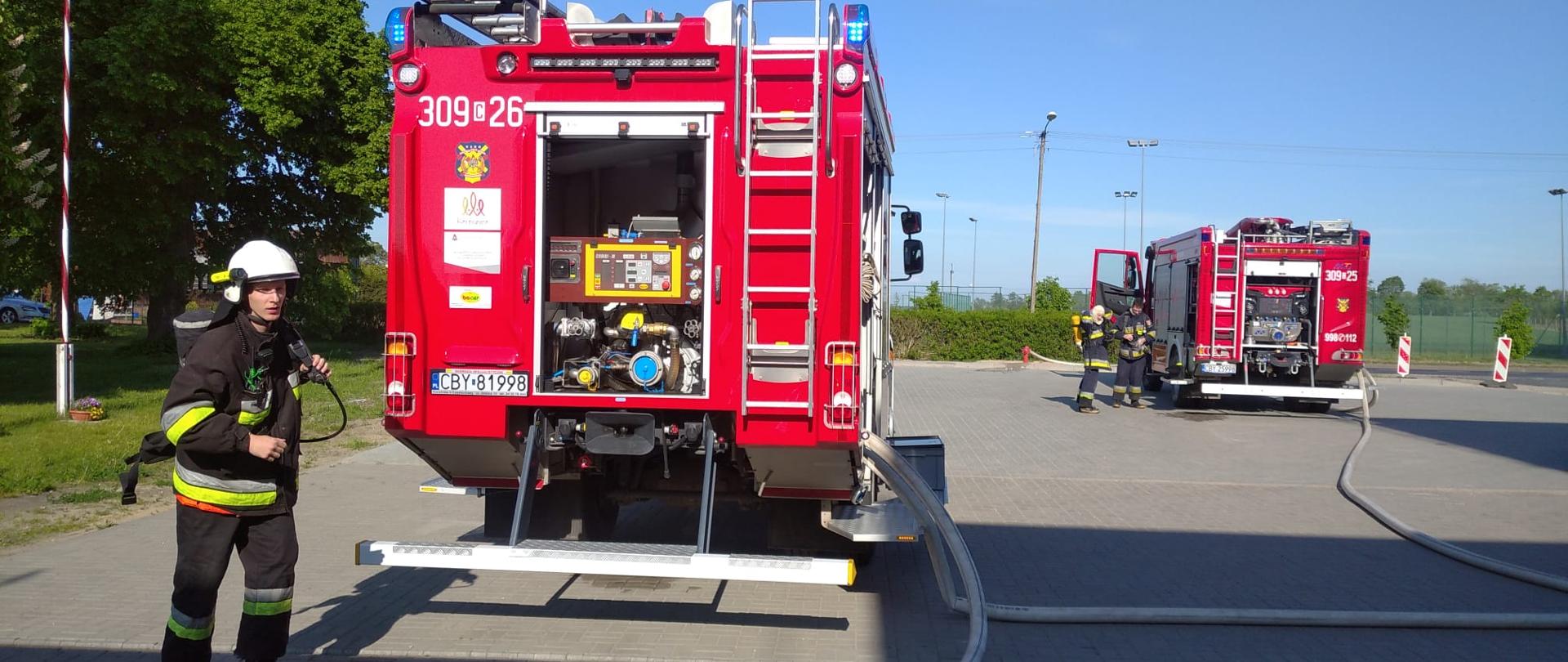 Dwa tyły ciężarowych pojazdów pożarniczych stojących na placu z szarej kostki brukowej, z lewej jeden strażak w granatowym ubraniu specjalnym i białym hełmie. Strażak ma na plecach aparat ochrony układu oddechowego. 