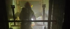 Na zdjęciu widać strażaka w pełnym umundurowaniu ochronnym, poruszającego się w zadymionym, słabo oświetlonym wnętrzu budynku. Postać jest widoczna od tyłu i oświetlona światłem latarki, co podkreśla gęsty dym unoszący się w pomieszczeniu. Strażak trzyma wąż gaśniczy prowadzący w głąb korytarza lub pomieszczenia. Wokół znajdują się elementy konstrukcyjne, takie jak metalowe barierki i rury, sugerujące ciasną, techniczną przestrzeń. Scena przedstawia akcję gaśniczą lub ćwiczenia w warunkach ograniczonej widoczności.