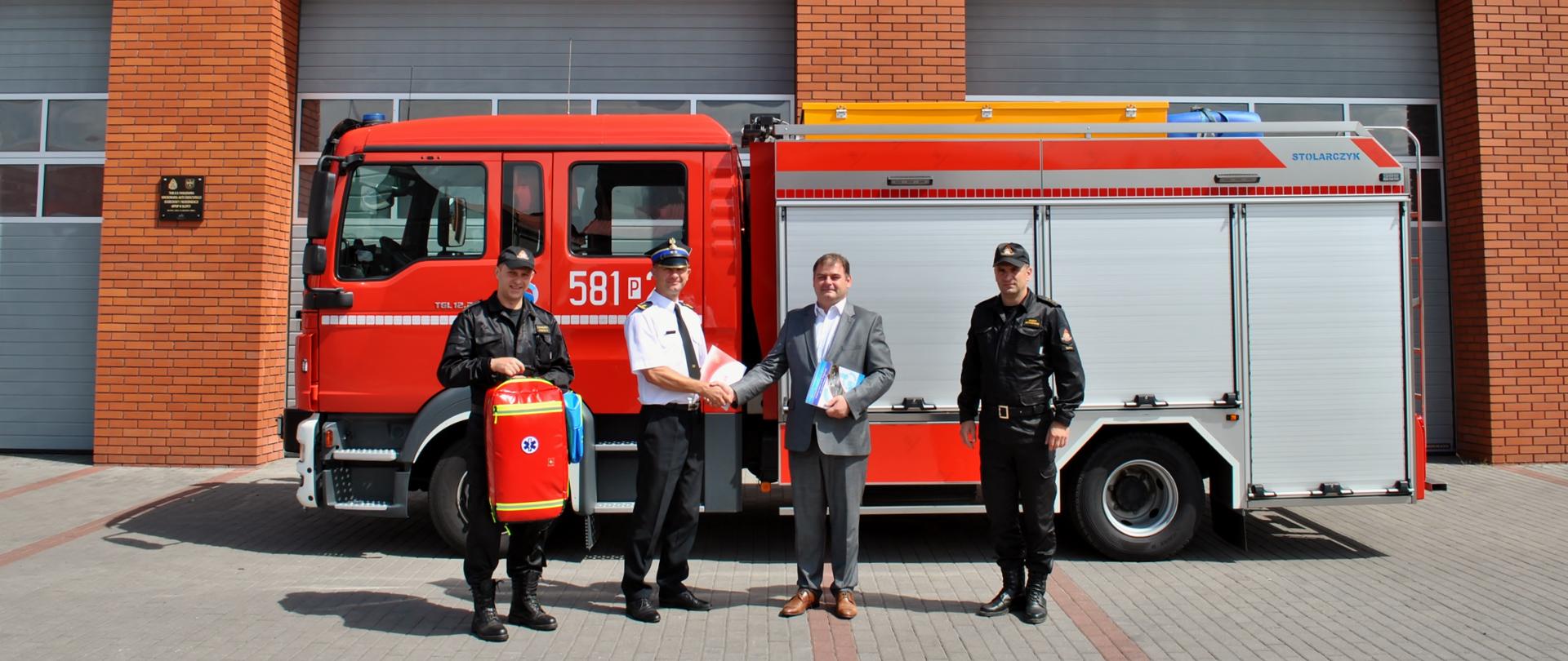 Przekazanie plecaka przez zastępce dyrektora WORD w szarym garniturze w obecności Komendanta Powiatowego oraz zastępcy Komendanta Powiatowego na tle samochodu strażackiego . Plecak trzyma strażak za nim budynek komendy z czerwonej cegły z napisem Państwowa Straż Pożarna