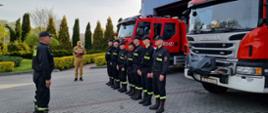 Zdjęcie przedstawia strażaków w umundurowaniu koszarowym. Strażacy podczas uroczystej zmiany służby stoją w dwóch szeregach naprzeciwko siebie. Po prawej stronie znajdują się dwa samochody specjalne. Na drugim planie widnieje strażak w ubraniu służbowym, który odczytuje rozkaz dzienny.