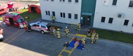 Wozy strażackie rozstawione na terenie zakładu podczas ćwiczeń, strażacy ćwiczą gaszenie pożaru w budynku oraz ratowanie osób poszkodowanych.
