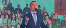 Mężczyzna w garniturze stoi z rozłożonymi rękami i przemawia przez mikrofon 