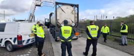 Grupa inspektorów transportu drogowego stoi wokół cysterny chemicznej podczas kontroli na terenie przemysłowym.