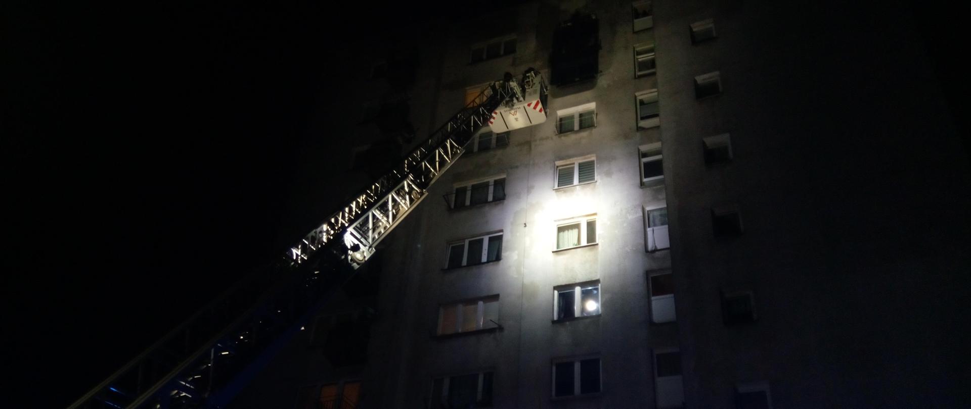 Zdjęcie przedstawia rozstawioną drabinę mechaniczną z koszem pod oknem na 9 piętrze wieżowca. Jest ciemno. Wieżowiec jest oświetlony. Nie widać ognia ani dymu.