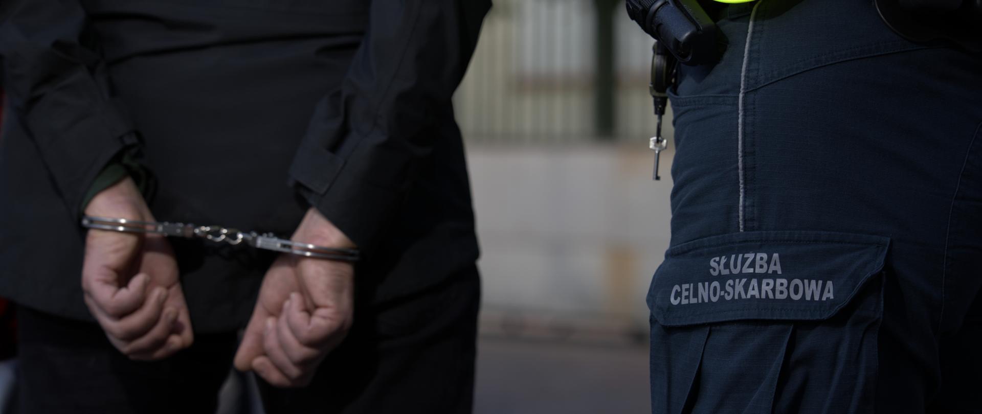 Na pierwszym planie widoczne są ręce osoby skute kajdankami trzymane za plecami. Obok znajduje się funkcjonariusz Służby Celno-Skarbowej w umundurowaniu z widocznym napisem „SŁUŻBA CELNO-SKARBOWA” oraz wyposażeniem służbowym.