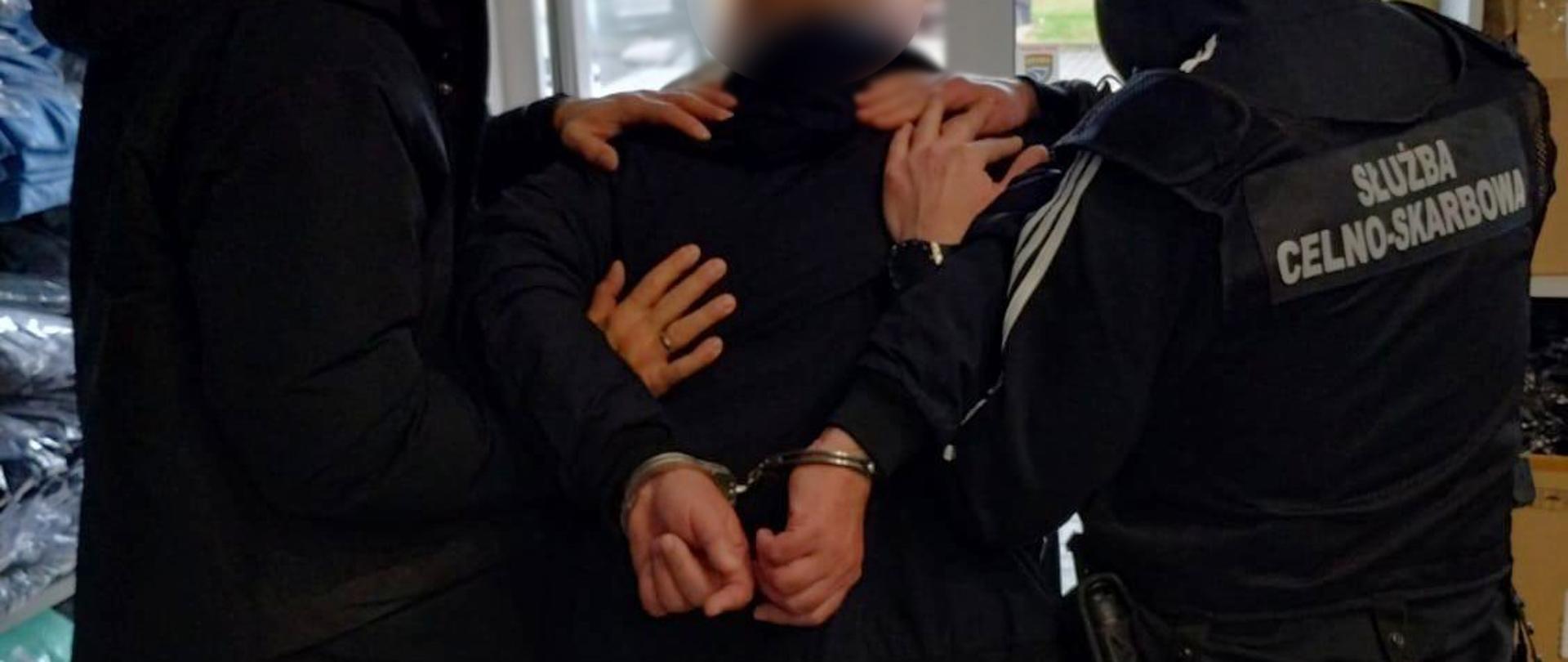 Funkcjonariusze w kamizelkach z napisem „Służba Celno-Skarbowa” zakładają kajdanki zatrzymywanej osobie.