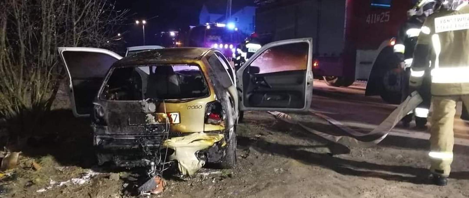Zdjęcie przedstawia tył spalonego samochodu osobowego marki Volkswagen. W pojeździe otwarte są drzwi kierowcy oraz pasażera. Po prawej stronie znajduje się dwóch strażaków w ubraniach specjalnych. W tle widnieją samochody ratowniczo-gaśnicze z włączoną alarmową sygnalizację świetlną. Zdjęcie zrobione jest w porze nocnej.