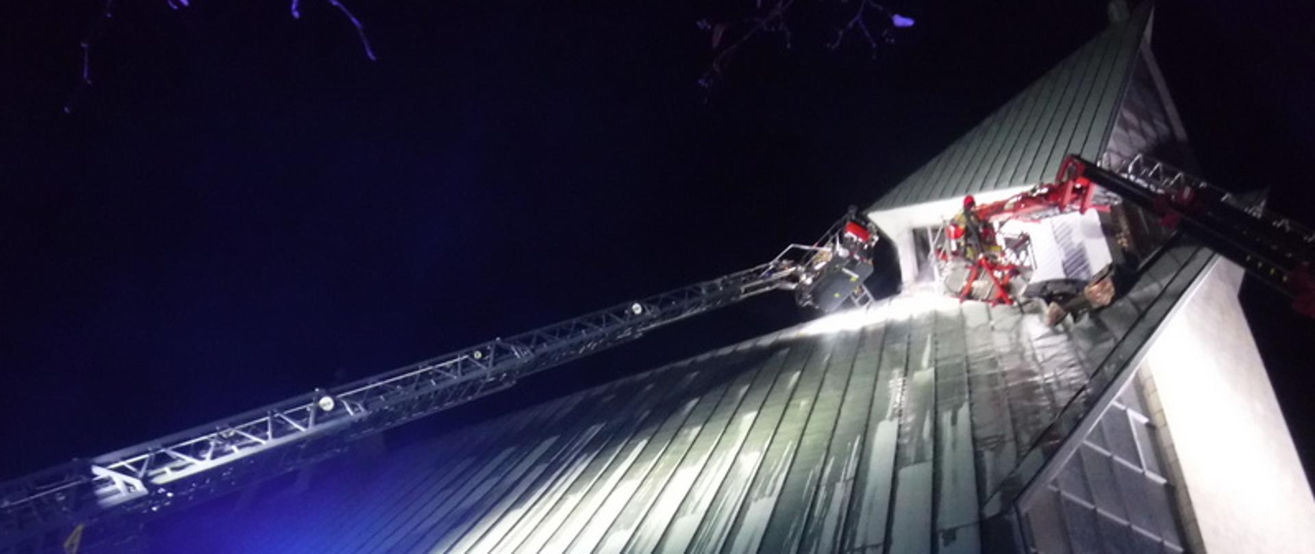 Zdjęcie przedstawia kościół nad którym widać rozłożone dwie drabiny hydrauliczne, w koszach tych drabin widać pracujących strażaków. Kościół i strażaków oświetlają lampy. Po lewej stronie zdjęcia widać odbijające się światło niebieskie . Jest pora nocna.