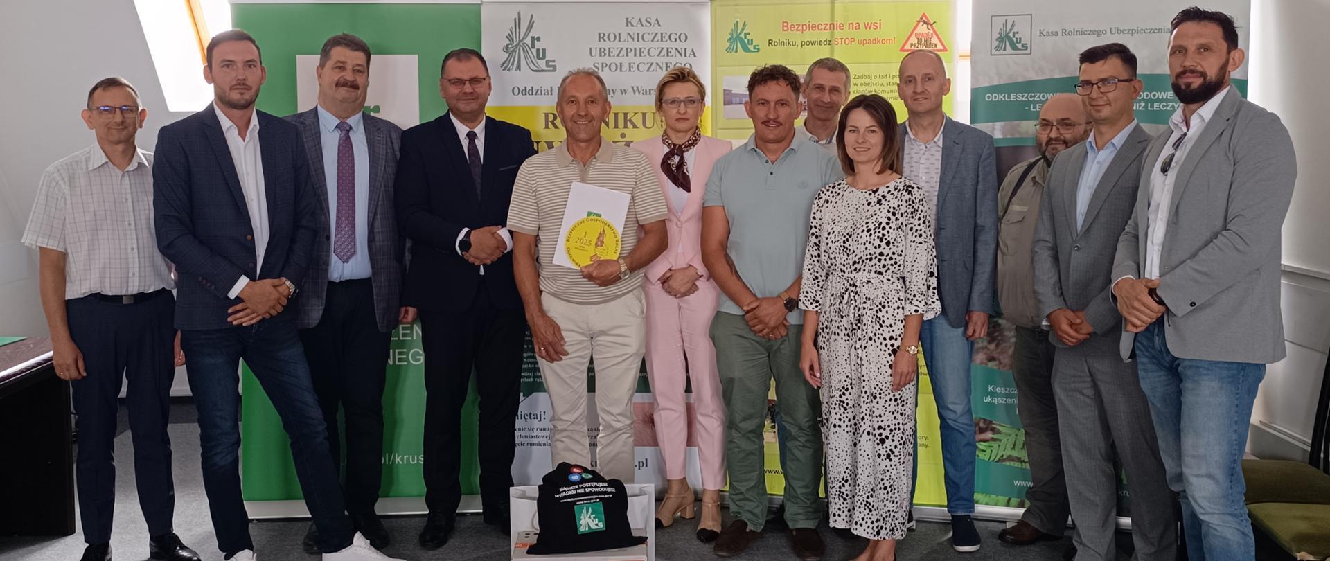 Grupa osób pozuje do zdjęcia na tle banerów prewencyjnych KRUS. Przed nimi stoją pudełka z nagrodami. Stojący w środku mężczyzna trzyma w ręce teczkę z logo konkursu.