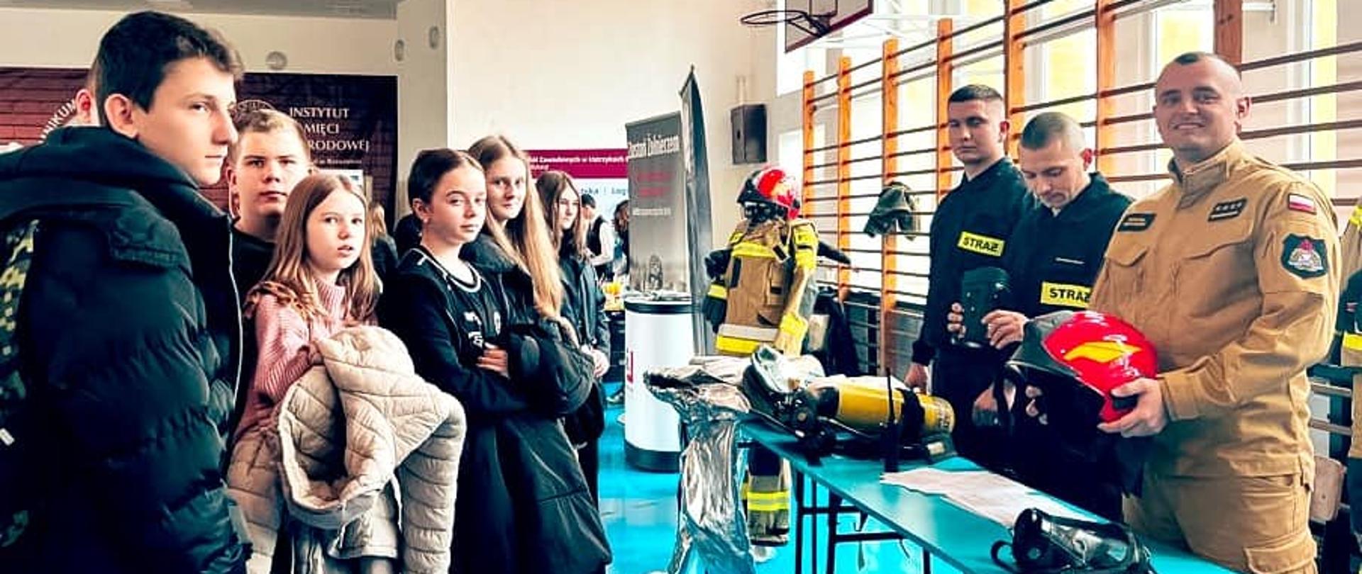 Na zdjęciu widać 3 strażaków oraz stoisko Komendy Powiatowej PSP w Ustrzykach Dolnych przy którym stoją uczniowie szkoły.