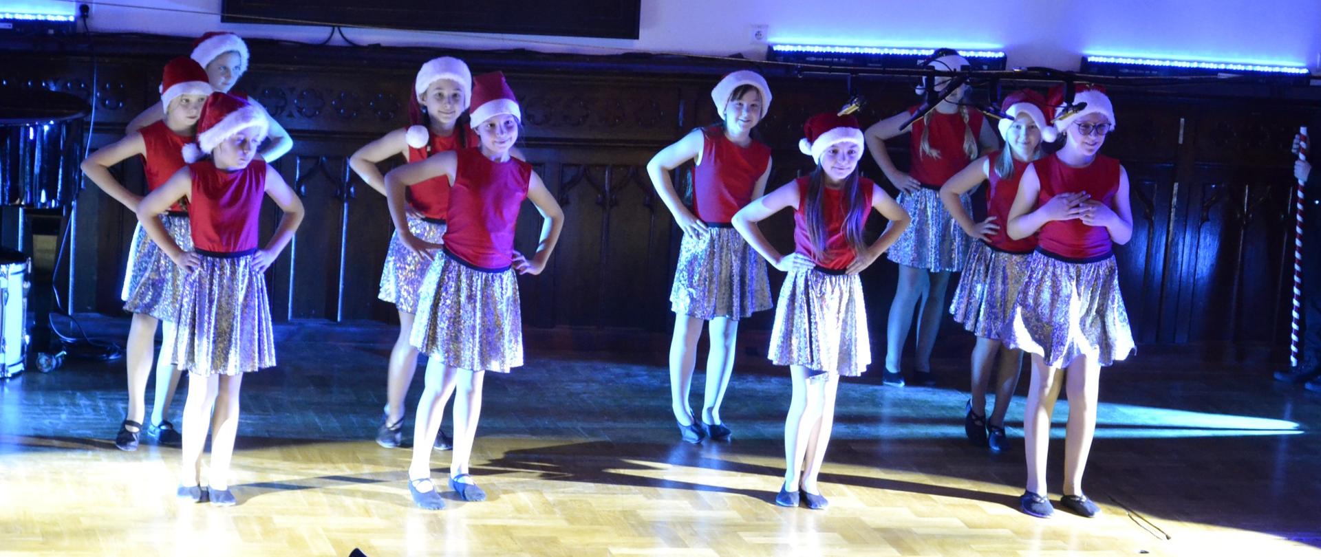 Grupa dziewczynek w świątecznych strojach – czerwonych bluzkach, błyszczących spódniczkach i czapkach Mikołaja – wykonuje taniec na scenie oświetlonej niebieskim i białym światłem. Mają energiczne pozy, z rękami na biodrach i uśmiechami na twarzach. W tle widać drewnianą boazerię i herb na ścianie, a na pierwszym planie stoją puste krzesła i pulpity na nuty.