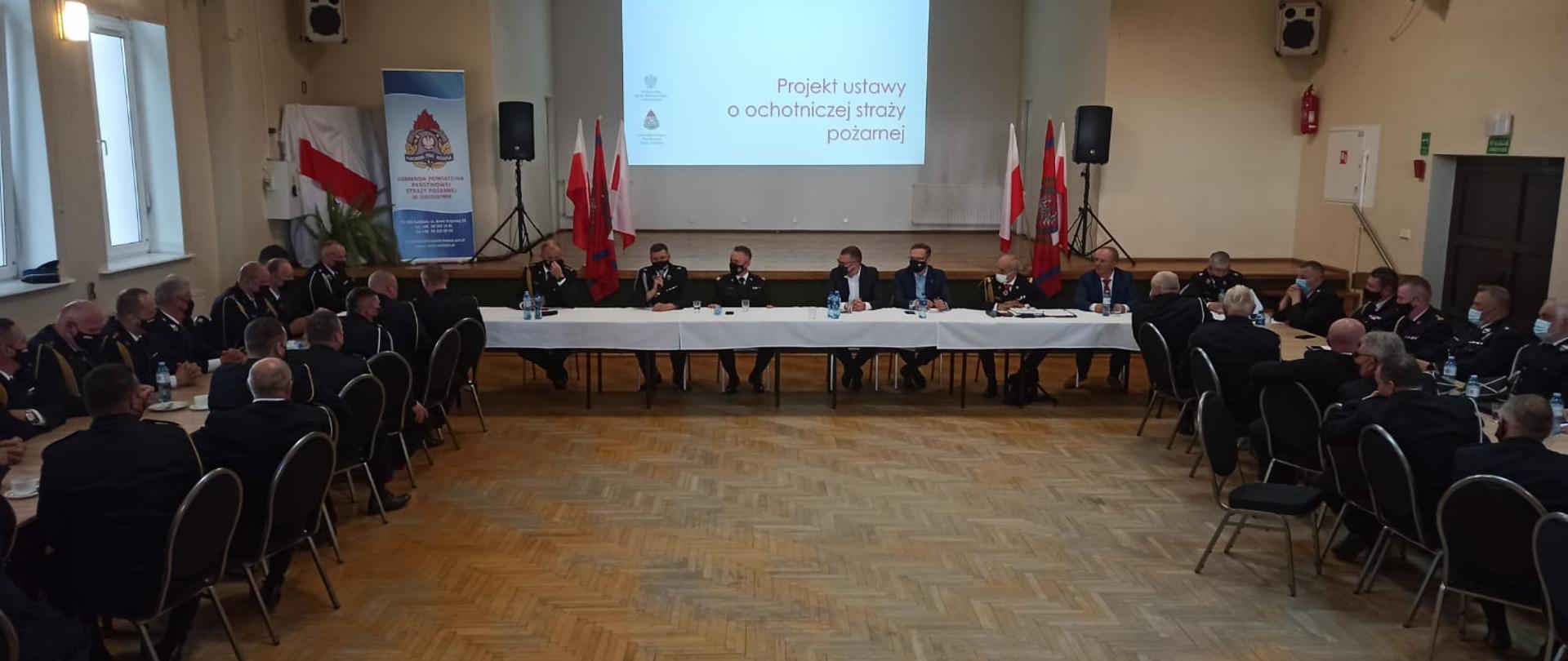 Zdjęcie przedstawia obradujących ludzi w dużej sali. Na środku sali przy stole siedzą: Sekretarz Stanu Ministerstwa Spraw Wewnętrznych i Administracji Paweł Szefernaker, Komendant Główny Państwowej Straży Pożarnej nadbryg. Andrzej Bartkowiak, Wojewoda Zachodniopomorski Zbigniew Bogucki, Zachodniopomorski Komendant Wojewódzki st. bryg. Jarosław Tomczyk. Po lewej stronie Komendanci Powiatowi/Miejscy województwa zachodniopomorskiego zaś po lewej przedstawiciele OSP.