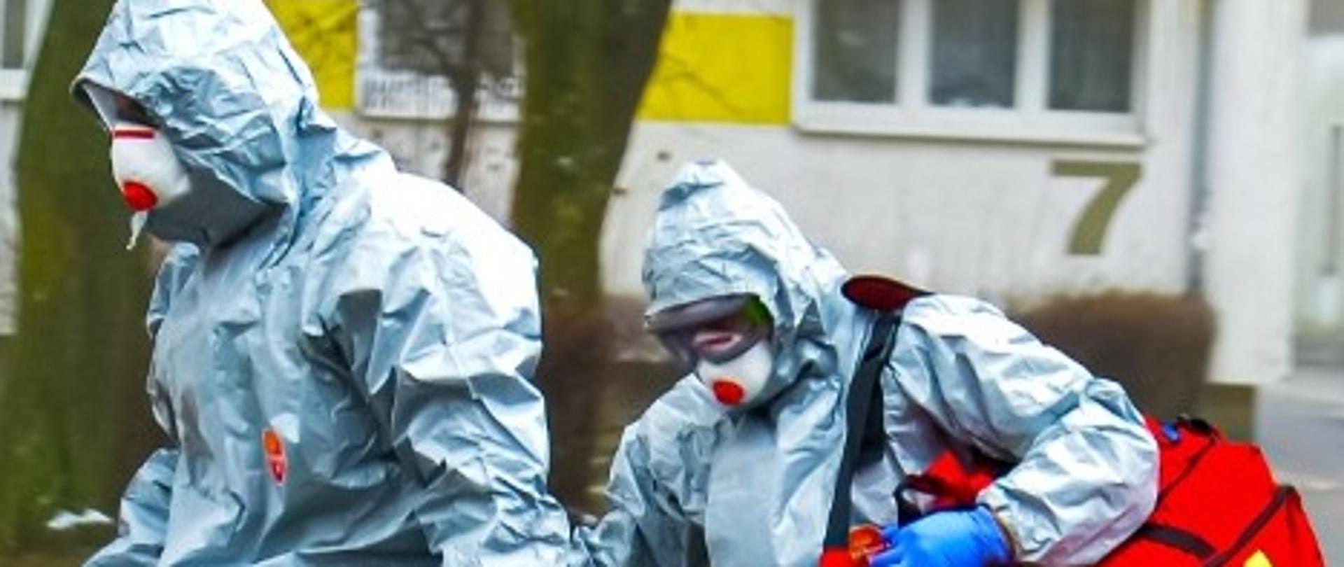 Dwóch ratowników z zestawem PSP R1 zabezpieczeni w ubrania ochrony biologicznej.