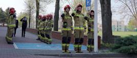 trzech strażaków stoi przy maszcie flagowym oddając honor fladze powiewającej na maszcie, po prawej stronie stoją cztery drzewa, plac wyłożony jest kostką brukową, za strażakami stojącymi przy maszcie widoczny namalowany na kostce znak określający miejsce parkingowe dla osób niepełnosprawnych w postaci dużego niebieskiego kwadratu z białą obwódką i symbolem osoby siedzącej na wózku inwalidzkim