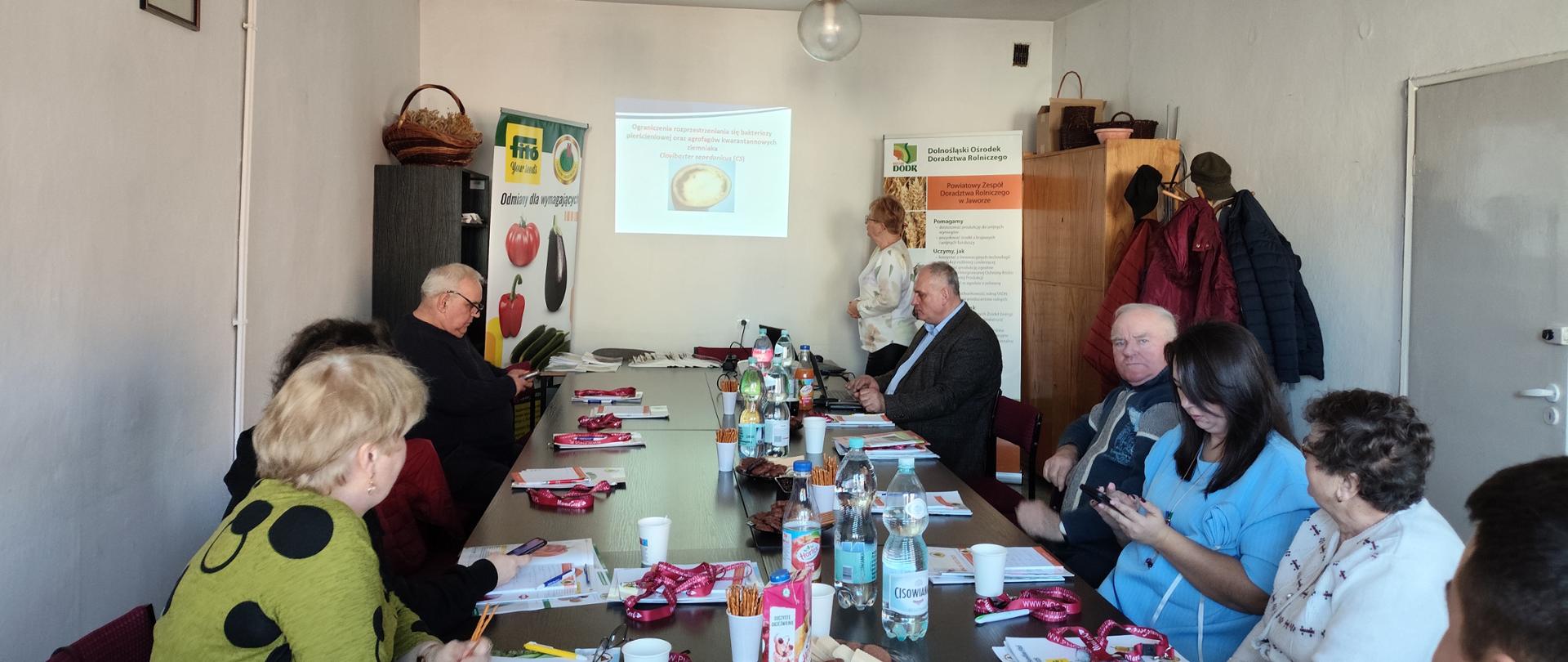 Uczestnicy spotkania siedzący przy stole. W tle pani kierownik Oddziału w Jaworze patrząca na wyświetloną prezentację dotyczącą Clavibacter sepedonicus